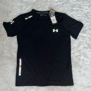 Svart Under Armour t-shirt - Svart t-shirt från Under Armour med reflekterande logga och tryck på bröstet och ärmarna. Tillverkad i lätt och snabbtorkande material, perfekt för träning. Klassisk passform med korta ärmar och rund hals. Snygg och sportig vibe!