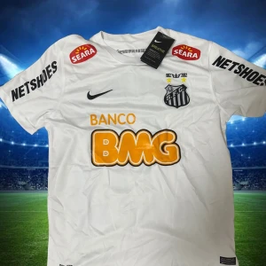 Santos Neymar Jr matchtröja Nike - Snygg vit Santos fotbollströja från Nike med Neymar Jr och nummer 11 på ryggen. Tröjan har korta ärmar, klubbmärke på bröstet och flera sponsorer tryckta i svart och orange. Materialet är lätt och andas, perfekt för träning eller match.