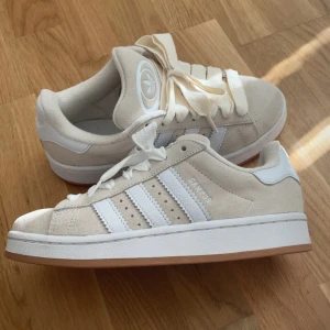 Beige Adidas Campus sneakers mocka - Helt nya Adidas Campus sneakers i beige mocka med vita detaljer och klassiska tre ränder på sidorna. Skorna har vit sula med gummidetalj och kommer med två par breda skosnören i vitt och offwhite. Perfekta för en clean och stilren look. Säljer för att har haft de länge och inte använt de. Skorna kom med offwhite skosnöre men säljs med ett par vita skosnöre. För mer information skriv privat/ pris kan diskuteras 💞
