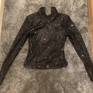 Svart paljettopp från NA-KD - Glittrig svart topp från NA-KD med lång ärm och hög krage. Toppens yta är täckt av paljetter som ger en riktigt festlig look. Stängs med knappar i nacken och har en figurnära passform. Perfekt för fester!!⭐️⭐️