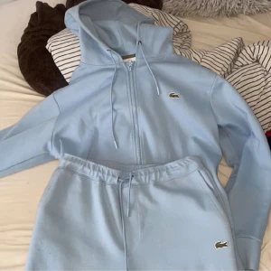 Ljusblå hoodie från Lacoste - Säljer en ljusblå tracksuit från Lacoste med dragkedja och huva. Tröjan har snörning vid huvan, långa ärmar och den klassiska krokodilloggan på bröstet. Perfekt för en chill och sportig look.