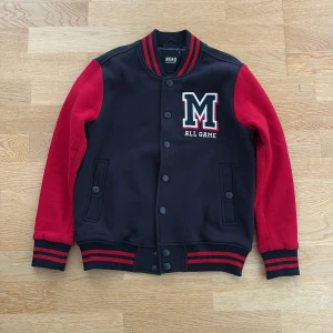 Svart och röd bomberjacka från MONO - Collegeinspirerad bomberjacka från MONO med svarta och röda färgblock, broderad M-logga och texten 'ALL GAME' på bröstet. Jackan har tryckknappar, randiga muddar och krage samt två fickor framtill. Perfekt för en sportig och avslappnad look.