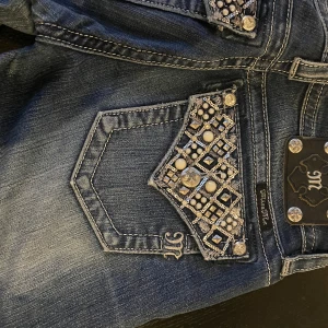 Miss Me skinny jeans med pärldetaljer - Snygga blå skinny jeans från Miss Me med coola bakfickor dekorerade med pärlor och nitar i silver. Jeansen har låg midja, klassisk femficksdesign och är tillverkade i stretchig denim för en tight passform. Perfekta för dig som gillar statement-detaljer!