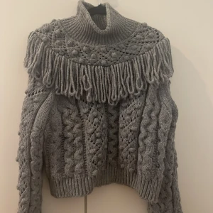 Grå stickad polotröja med fransar Zara - Säljer en chunky grå stickad polotröja från Zara med snygga flätmönster och långa fransar över axlarna. Tröjan har hög krage, lång ärm och en loose fit som ger en cool och mysig vibe. Perfekt för kalla dagar när du vill sticka ut lite extra.
