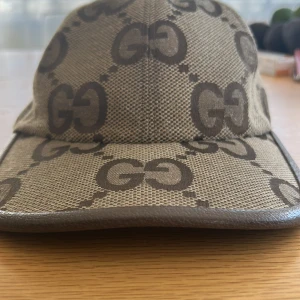 Gucci keps med GG-mönster - Snygg keps från Gucci i beige och brunt med det klassiska GG-monogrammet över hela kepsen. Justerbar rem baktill i brunt skinn och böjd skärm. Tillverkad i canvas med läderdetaljer för en lyxig känsla. Perfekt för dig som vill sticka ut med en ikonisk accessoar.