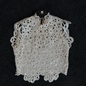 Beige spetsblus med paljetter från Maje - Superfin beige blus från Maje med virkad spets i blommigt mönster och glittrande paljetter. Ärmlös modell med hög hals och snygg öppning bak. Blusen har ett innerfoder i mjuk polyesterblandning för extra komfort. Perfekt för dig som gillar unika detaljer!