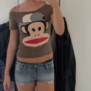 Brun Paul Frank offshoulder t-shirt - Säljer en brun offshoulder t-shirt från Paul Frank med stort tryck av Julius-apan på framsidan. T-shirten är tight och har korta ärmar, perfekt för dig som gillar en lekfull och trendig stil.