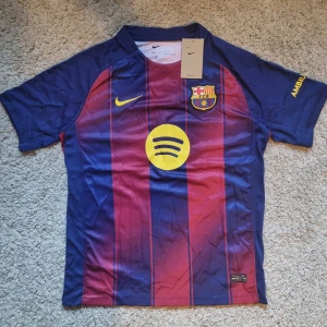 FC Barcelona 26 tröja - FC Barcelona matchtröja från Nike i storlek M. Klassisk design med blå och vinröda vertikala ränder, klubbmärke på bröstet och gula detaljer som Nike-logga och sponsortryck. Tillverkad i lätt och ventilerande Dri-FIT-material, perfekt för fotboll eller supporterliv.
