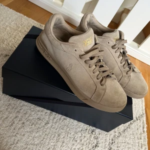 Polo Ralph Lauren skor - Stilrena sneakers i beige mocka från Polo Ralph Lauren med klassisk låg profil och rund tå. Skorna har snörning, diskret logga på sidan och guldiga Polo-detaljer på plös och sula. Perfekta för en clean och avslappnad look.