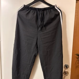 Svarta mjukisbyxor med vita ränder - Svarta mjukisbyxor med vita dubbla ränder längs sidorna. Byxorna har elastisk midja med snörning och en avslappnad passform. Perfekta för chill dagar eller när du vill ha det bekvämt. Materialet känns mjukt och lätt.