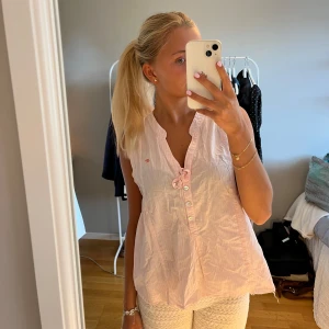 Randig ärmlös blus med rosett - Söt ärmlös blus i vitt och ljusrosa med tunna ränder, knappar framtill och en liten rosett vid halsringningen. Liten broderad logga på bröstet. Storlek L men passar mig som har S