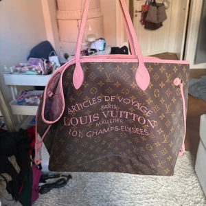 Vintage Louis Vuitton shoppingväska med rosa detaljer - Louis Vuitton shoppingväska i klassiskt brunt monogram med rosa handtag och kanter. Insidan är fodrad med blommigt rosa tyg och har en innerficka med dragkedja. Perfekt för dig som vill ha en lyxig och rymlig väska med unik touch.