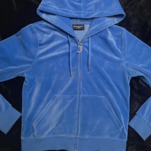 Blå velour hoodie från Juicy Couture - Juicy Couture hoodie i blå velour med dragkedja och huva. Klassisk J-dragkedja, långa ärmar och känguruficka framtill. Inga defekter och säljer pga kommer inte till användning. Köpt på plick, därav bilderna lånade ifrån.