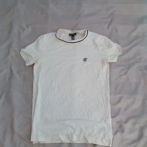 Vit t-shirt från SikSilk med svart kant - Vit t-shirt från SikSilk med svart broderad logga på bröstet och svart kant runt halsen. Klassisk passform och korta ärmar. Tillverkad i mjuk bomull som känns skön mot huden. Perfekt för en clean och stilren look.