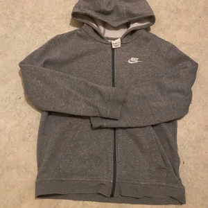 Grå hoodie från Nike med dragkedja - Snygg grå hoodie från Nike med vit logga på bröstet. Tröjan har huva, hel dragkedja och två fickor framtill. Tillverkad i mjuk bomullsblandning och har långa ärmar. Perfekt för chill eller träning.