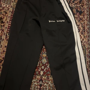 Palm Angels svarta mjukisbyxor med ränder - Svarta mjukisbyxor från Palm Angels med vita sidoränder och broderad logga på benet. Byxorna har elastisk midja med dragsko och sportig vibe. Perfekta för en chill och trendig streetwear-look.