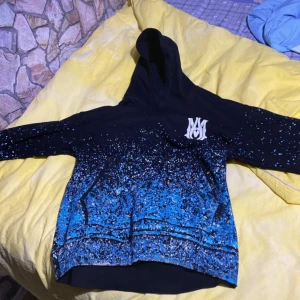 AMIRI hoodie svart/blå stänk  - Säljer en fet hoodie från AMIRI i svart med blått och vitt stänkmönster. Stor logga på ryggen och mindre tryck fram. Tröjan har huva, känguruficka och är gjord i 100% bomull. Perfekt för dig som vill sticka ut pris kan diskuteras.