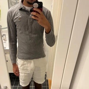 Grå långärmad pikétröja Polo Ralph Lauren - Snygg grå långärmad pikétröja från Polo Ralph Lauren i custom slim fit. Klassisk krage, knäppning med tre knappar och brunt tyg på insidan av knappslån. Ikonisk broderad logga på bröstet. Perfekt för en clean och stilren look.