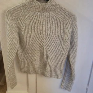 Grå ribbad stickad tröja - Säljer en beige stickad tröja med ribbat mönster och hög hals. Tröjan har långa ärmar och ett snyggt strukturerat mönster som ger en modern vibe. Perfekt för lager-på-lager och passar till både jeans och kjol.