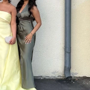 Olivgrön långklänning med rosdetalj - Supersnygg olivgrön långklänning i glansigt satinliknande material. Klänningen har halterneck, v-ringning och en rosdetalj framtill. Perfekt som fest eller bal klänning 