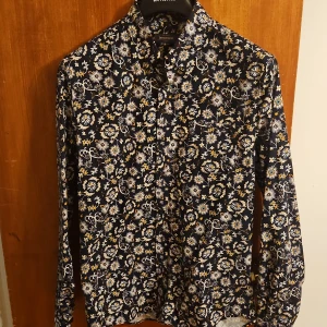 Blommig skjorta från Dressmann L - Snygg långärmad skjorta från Dressmann i slim fit-modell. Skjortan är marinblå med blommigt mönster i vitt, gult och blått. Perfekt för dig som vill sticka ut med en färgglad och trendig look. Knappar framtill och klassisk krage.