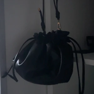 Svart bucket bag med snörning - Säljer en svart bucket bag i glansigt skinnimitation med snörning upptill och guldfärgade metalldetaljer. Väskan har justerbar axelrem och en cool, modern look som passar perfekt till streetstyle eller när du vill sticka ut lite extra.