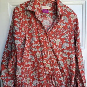 Röd blommig skjorta från Morris Lady - Supersnygg röd skjorta från Morris Lady i samarbete med Liberty, täckt av blommigt mönster i vitt, gult och grönt. Skjortan har klassisk krage, knäppning framtill och långa ärmar. Perfekt för dig som vill sticka ut med färg och mönster.