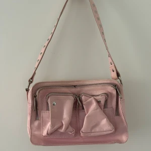 Ljusrosa axelväska från Nunoo - Säljer en ljusrosa axelväska i skinn från Nunoo, den har flera fickor framtill och silvriga detaljer. Väskan har ett axelband med nitar men ett långt band ingår. Superfin att ha till en simpel outfit! Väskan är i bra skick! 