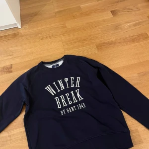 Mörkblå sweatshirt från Gant - Snygg mörkblå sweatshirt från Gant med vit text 'Winter Break by Gant 1949' på bröstet. Tröjan har rund halsringning, långa ärmar och ribbade muddar. Perfekt för dig som gillar klassisk och sportig stil.