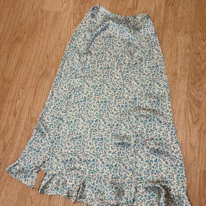 Blommig långkjol från Zara XS - Supersöt satin långkjol från Zara i storlek XS med blommigt mönster i blått och orange på en ljus bakgrund. Kjolen har volangkant nedtill och dragkedja i sidan. Perfekt för dig som gillar romantisk och feminin stil.