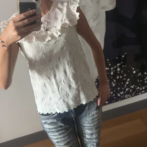 Vit blus med volang och spets - Supersöt vit blus med volang runt halsen och ärmlös design. Blusen har ett spetsliknande mönster och vågig kant nertill, perfekt för en fräsch och somrig look. Lätt att matcha med både jeans och kjol.