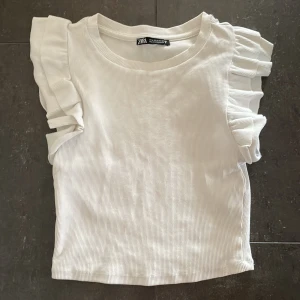 Vit  topp med volangärmar Zara - Superfin vit ribbad topp från Zara med volangdetaljer på axlarna. Toppen är ärmlös och har en rund halsringning. Perfekt för dig som gillar en söt och trendig stil med extra detaljer på axlarna.