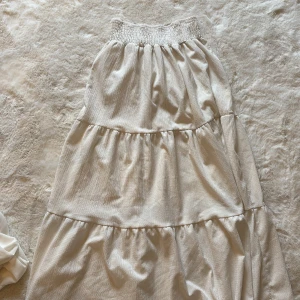 Vit långkjol från 157 - Supersöt vit långkjol från 157 i storlek S. Kjolen har flera volanger och ett smockat midjeband som gör den extra bekväm. Materialet är lätt och har en diskret struktur som ger en snygg touch. Perfekt för dig som gillar flowiga och luftiga plagg.