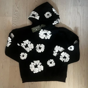 Svart hoodie med vita blommor Denim Tears - Svart hoodie från Denim Tears med stora vita blommor tryckta över hela tröjan. Tröjan har huva och en relaxed passform, perfekt för dig som gillar streetwear med unik design. Materialet är mjuk bomull och trycket ger en cool kontrast mot det svarta tyget.