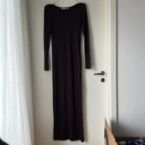 Mörkbrun långklänning från Aware - Långärmad, mörkbrun långklänning från Aware i figurnära modell. Klänningen har rund halsringning och är tillverkad i ett mjukt, stretchigt material som sitter snyggt på kroppen. Perfekt för dig som gillar en stilren och minimalistisk look.