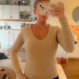 Beige ribbad stickad tröja - Snygg beige stickad tröja med ribbad struktur och djup rundad halsringning. Tröjan har långa ärmar och sitter normalt på kroppen, perfekt för dig som gillar stilrena och enkla plagg med lite edge. Passar till jeans eller kjol och är superenkel att matcha.