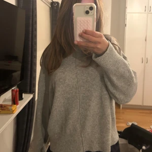 Grå stickad polotröja från Stories - Mysig grå stickad polotröja från Stories i storlek S. Tröjan har en hög krage, långärmade och loose passform med ribbade muddar och nederkant. Perfekt för kyliga dagar och enkel att styla med jeans eller kjol. Snygg söm framtill som ger en cool detalj.