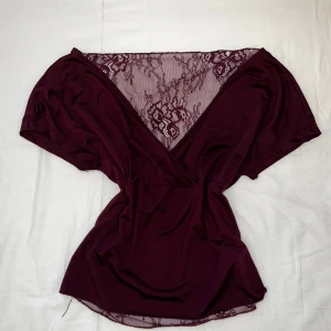 Vinröd kort omlotttopp med spets - Supersnygg vinröd topp med omlott-design och djup v-ringning. Toppen har breda korta ärmar och detaljer i transparent spets där bak, vilket ger en extra touch av elegans. 😝