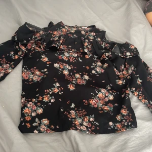 Svart blommig blus med volang - Supersöt svart blus med blommigt mönster i rött, grönt och beige. Blusen har volangdetaljer upptill och långa ärmar med resår vid handlederna. Öppna axlar. Perfekt för dig som gillar romantisk stil och vill sticka ut lite extra.