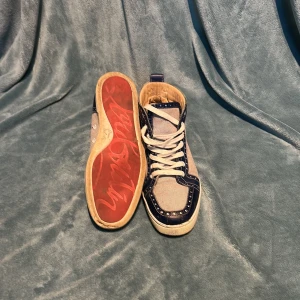 Louboutin sneakers blå/vit, stl 42/43 - Snygga high-top sneakers från Louboutin i blått och vitt med coola nitar och klassisk röd sula. Skorna har vita snören, detaljer i skinn och textil, samt Louboutin-logga på sidan.  Dessa är en såpas gammal moddel, var tvungen att byta sula pågrund av att den gick sönder så båda är bytta till silikon underlag. Skriv om ni har några frågor 