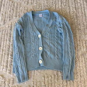 Ljusblå kofta från Lindex med knappar - Säljer en ljusblå kofta från Lindex med snyggt flätstickat mönster och stora vita knappar framtill. Koftan har v-ringad hals och långa ärmar med ribbade muddar. Perfekt att slänga över en t-shirt när det är lite kyligt ute.