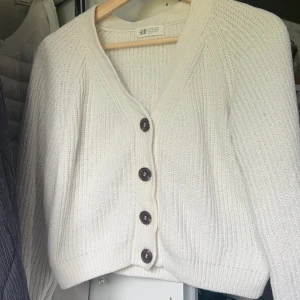 Vit stickad kofta från H&M - Supermysig vit stickad kofta från H&M med stora mörka knappar framtill. Koftan har v-ringning, lång ärm och en lite loose passform. Perfekt att slänga på sig när det är kyligt ute. Enkel att matcha med jeans eller kjol.