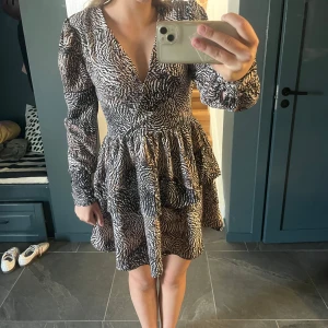 Zebramönstrad kortklänning med v-ringning - Supersnygg kort klänning med zebramönster i svart och beige. Klänningen har v-ringning, långa ärmar och markerad midja med volangig kjol. Perfekt för dig som vill sticka ut med ett djurmönster och gillar trendiga detaljer.