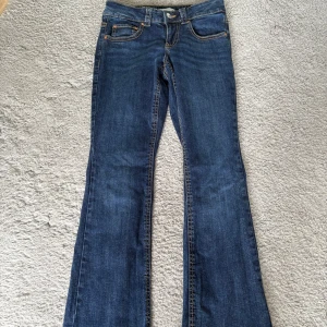 Blå bootcut jeans från Gina Tricot - Snygga blå jeans från Gina Tricot med bootcut-modell och klassiska kontrastsömmar. Jeansen har normalhög midja, fem fickor och en snygg detalj med ficklock bak. Perfekta för dig som gillar en tidlös och trendig look.