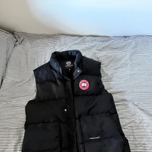 Svart dunväst från Canada Goose - Säljer en svart dunväst från Canada Goose med klassisk logga på bröstet. Västen har hög krage, stora fickor framtill och stängs med både dragkedja och tryckknappar. Perfekt för lager-på-lager och riktigt snygg till streetwear. Den är helt ny fick den present av en vän på min födelsedag och Scan finns med också! Priset kan diskuteras 