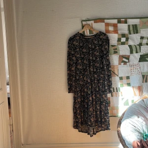 Svart blommig långklänning med knappar - Svart klänning med blommigt mönster i beige och gult. Klänningen har rund halsringning, långa ärmar och knapp i nacken. Materialet känns mjukt och lätt! 