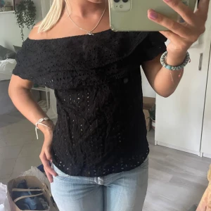 Svart offshoulder blus med spets - Säljer en svart offshoulder blus med snygga spetsdetaljer och hålmönster. Blusen har en bred volang upptill som ger en cool och feminin vibe. Perfekt för dig som gillar att sticka ut med detaljer och älskar svart. Passar till jeans eller kjol för en trendig look.