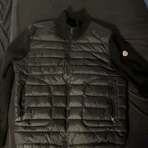 Svart quiltad pufferjacka från Moncler - Snygg svart cardigan från Moncler med quiltad framsida och släta ärmar. Jackan har dragkedja framtill, två sidofickor med dragkedja och Moncler-logga på ena ärmen. Perfekt för dig som gillar stilren design och exklusiva märken. Storlek M och i mycket bra skick.