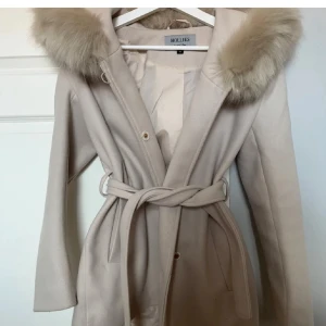Beige kappa med fuskpäls från Hollies - Snygg beige kappa från Hollies, fick en liten fläck inuti jackan men inget som syns utifrån. Prisförslag går att diskutera 