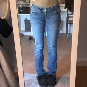 True Religion ljusblå bootcut jeans - Säljer ett par ikoniska ljusblå jeans från True Religion med bootcut passform och snygga detaljerade bakfickor. Jeansen har slitning och hål på ena låret för en edgy look. Klassisk femficksmodell och låg midja. Perfekta för dig som gillar streetstyle. Köpta på second hand.